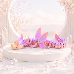 Könnte beinhalten: Eine bewegliche Drachenfigur in Rosa, Lila und Gold ruht auf einem weißen Podest. Der Drache hat große, schillernde Flügel und einen eingerollten Schwanz. Der Hintergrund zeigt zarte rosa Blüten und Kristalle.
