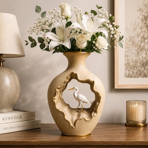 May include: A beige ceramic vase with a textured finish and a central hollow opening containing a small white heron figurine. The vase is filled with a bouquet of white lilies, white roses, and sprigs of baby's breath, creating a floral arrangement.