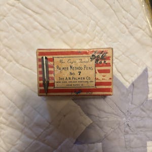 May include: A rectangular box with red and white stripes containing a metal pen nib. The label reads: New Engine Turned PALMER METHOD PENS No. 7 THE A.N.PALMER CO. NEW YORK CHICAGO PORTLAND ORE CEDAR RAPIDS IA. The box shows signs of wear and age.