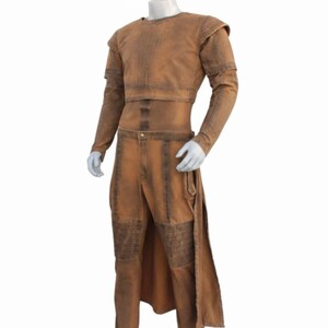 Op de afbeelding: Een bruin kostuum uitgestald op een mannequin. De outfit bevat een top met lange mouwen en schoudervullingen, een bijpassende broek en een lang rokachtig paneel. De stof heeft een verweerde uitstraling.