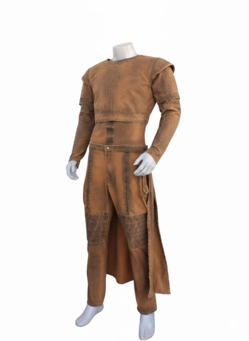 Op de afbeelding: Een bruin kostuum uitgestald op een mannequin. De outfit bevat een top met lange mouwen en schoudervullingen, een bijpassende broek en een lang rokachtig paneel. De stof heeft een verweerde uitstraling.
