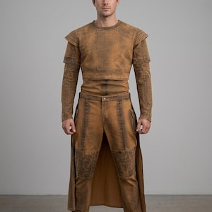 Op de afbeelding: Een persoon draagt een tweedelige outfit in een verweerde bruine kleur. De top is een shirt met lange mouwen en schouderaccenten. De broek is bijpassend bruin met kniebeschermers en zijzakken. De outfit wordt gecompleteerd met bruine laarzen.