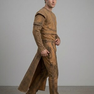 Op de afbeelding: Een bruine outfit bestaande uit een top met lange mouwen, een korte jas, een lange broek en een lange jas. De outfit heeft een distressed, getextureerde uitstraling. De broek heeft kniebescherming. De persoon draagt bruine laarzen.