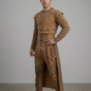 Op de afbeelding: Een tweedelige outfit in een verbleekte bruine kleur. De top is een cropped shirt met lange mouwen en schouderaccenten. De broek heeft een hoge taille met een lange, split rok overlay en kniebeschermers. Het model draagt bruine laarzen.