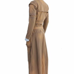Op de afbeelding: Een bruin, lange mouwen outfit met een bijpassende lange rok. De outfit heeft meerdere zakken, een riem en schouderdetails. Het kledingstuk wordt op een mannequin gepresentd tegen een witte achtergrond.