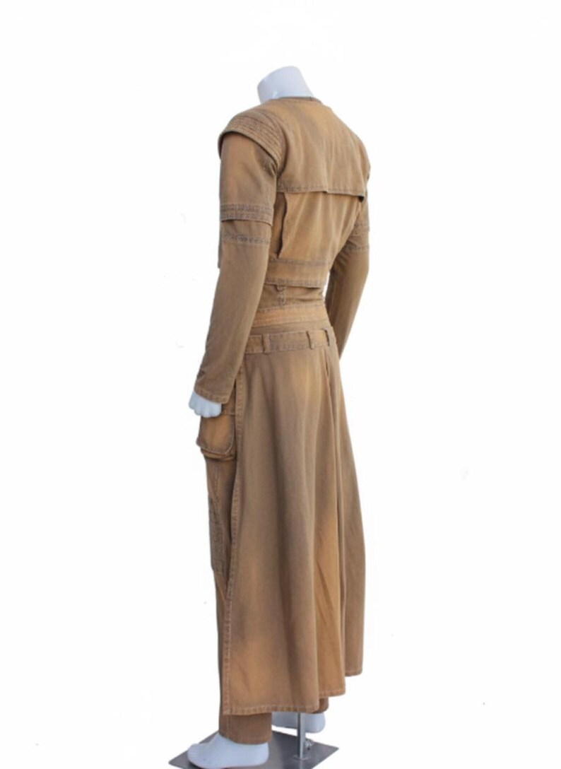 Op de afbeelding: Een bruin, lange mouwen outfit met een bijpassende lange rok. De outfit heeft meerdere zakken, een riem en schouderdetails. Het kledingstuk wordt op een mannequin gepresentd tegen een witte achtergrond.