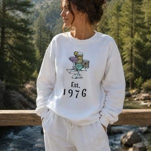 Puede incluir: Una sudadera blanca de cuello redondo con una ilustración gráfica de cuatro cócteles diferentes en varios vasos. Debajo de la ilustración de los cócteles, el texto dice Est. 1976 en una fuente serif negra. La sudadera tiene mangas largas.
