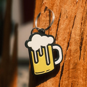 Könnte beinhalten: Ein Schlüsselanhänger aus Gummi in Form eines Bierkrugs mit gelbem Körper, weißem Schaumkrone und schwarzer Umrandung. Das Design zeigt einen weißen Henkel und weißen Schaum, der an den Seiten des Krugs herabläuft. Ein silberner Metallring ist befestigt.