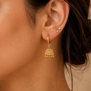 May include: Gold-coloured hoop earrings with a bell-shaped charm. The earrings feature intricate filigree detailing and small dangling beads. The model also has two small stud earrings in the ear.