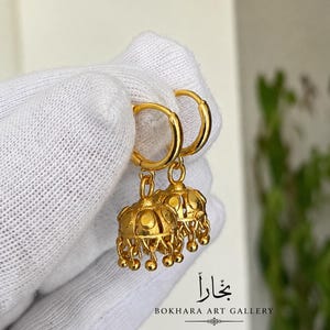 Könnte beinhalten: Ein Paar goldene Creolen mit glockenförmigen Jhumka-Anhängern. Jeder Anhänger hat einen glatten, abgerundeten oberen Teil mit kleinen, baumelnden goldenen Kugeln entlang der Unterkante. Die Ohrringe haben eine helle, reflektierende Oberfläche.