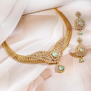 May include: A gold-toned necklace and matching pair of earrings featuring light blue rectangular and teardrop-shaped stones. The jewelry set is embellished with numerous small, clear, sparkling stones arranged in intricate, radiating patterns.