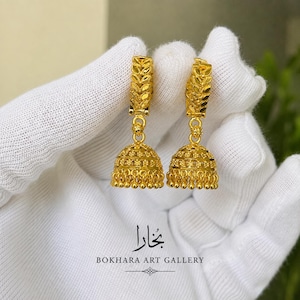 May include: A pair of gold jhumka earrings featuring a textured leaf-patterned hoop top and a bell-shaped drop with intricate bead detailing. The earrings are displayed against a white fabric background with the text BOKHARA ART GALLERY.