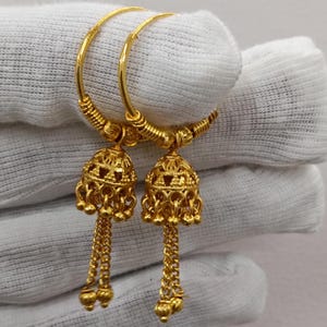 May include: A pair of gold hoop earrings featuring dangling bell-shaped jhumka charms. Each charm has intricate cut-out patterns and is finished with two delicate gold chains ending in small spherical beads, creating a detailed metallic design.