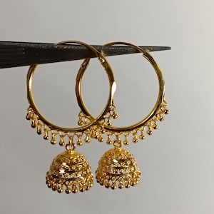 Könnte beinhalten: Ein Paar goldene Creolen, bestehend aus einem kreisförmigen Ring mit kleinen hängenden Perlen am unteren Rand. Jeder Reifen trägt einen glockenförmigen Jhumka-Anhänger mit filigraner Metallarbeit und einer Reihe winziger Goldkugeln am Boden.