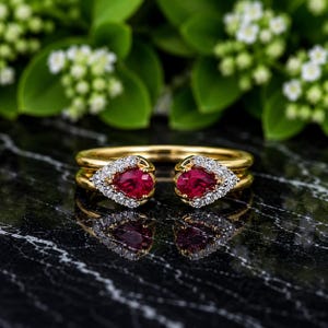 Puede incluir: Un anillo abierto de oro con dos rubíes rojos en forma de pera, cada uno rodeado por un halo de pequeños diamantes blancos. La banda está hecha de oro amarillo pulido, creando un diseño bypass que deja un espacio entre los dos engastes.