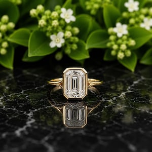May include: A gold ring featuring a large, rectangular emerald-cut clear gemstone set in a smooth bezel setting. The band has a split shank design that tapers toward the center stone, creating a clean, polished look with a reflective metallic finish.