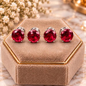 May include: Four round, faceted, deep red gemstone stud earrings set in silver-toned metal prongs. Each earring features a brilliant cut, creating a reflective surface. The studs are arranged in a horizontal row on a beige velvet display.