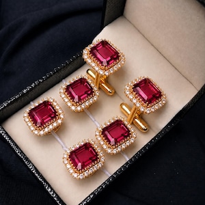 May include: A set of four gold-toned cufflinks and a matching stud, each featuring a rectangular, deep red faceted stone surrounded by a border of small, clear, sparkling stones. The pieces are arranged inside a black, textured display box.