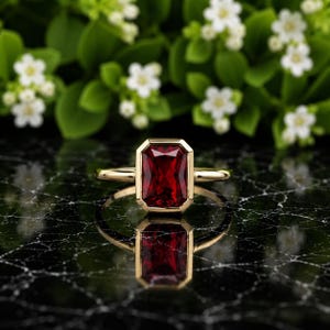 May include: A gold ring featuring a large, rectangular, emerald-cut deep red gemstone set in a smooth, polished bezel setting. The ring is centered, showcasing a vibrant red stone with sharp, clean lines and a reflective, metallic gold band.
