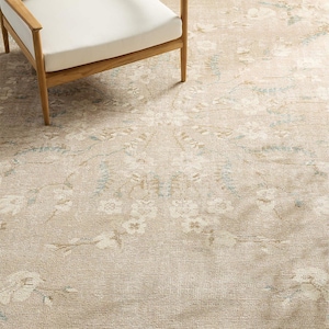 May include: A large area rug featuring a beige base with a faded floral pattern in white and soft blue tones. The rug has a textured, woven appearance and is partially covered by a wooden chair with a white seat cushion and light wood frame.