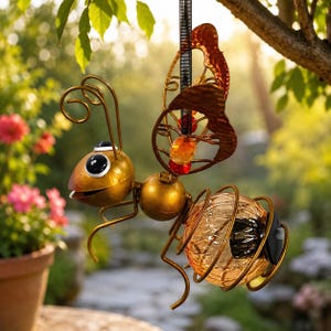 May include: A hanging metal bee ornament featuring a gold-toned head with large black and white eyes, wire antennae, and wings accented with red and orange beads. The body consists of a textured amber glass sphere encased in a gold wire frame.