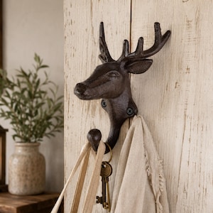 Könnte beinhalten: Ein dunkelbrauner Wandhaken aus Gusseisen in Form eines Hirschkopfes mit Geweih. Der Haken ist mit zwei sichtbaren Schrauben an einer hellen Holzoberfläche befestigt. Eine beigefarbene Stofftasche und ein Schlüsselbund hängen am unteren Haken.