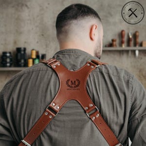 May include: A tan leather harness with an X-shaped back panel featuring a debossed laurel wreath emblem, the letter M, and the name Michael. The harness is constructed with silver-toned metal rivets and adjustable straps worn over a gray shirt.