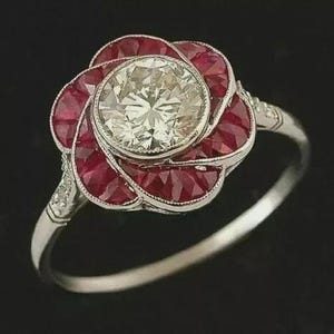 May include: A silver ring featuring a flower design with a large, round, clear center stone and surrounding red stones. The ring's band is thin and has small accent stones on the sides of the flower.
