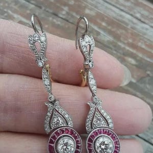 May include: Elegant drop earrings featuring a central round diamond surrounded by a halo of small red gemstones. The earrings have a silver-tone metal setting with intricate detailing and additional small diamonds. The design includes a decorative top and a long, slender drop.