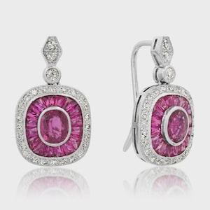 May include: A pair of silver drop earrings with a square design. Each earring features a central oval of pink gemstones surrounded by a ring of pink gemstones and a border of small white gemstones. The top has a small diamond-shaped design.