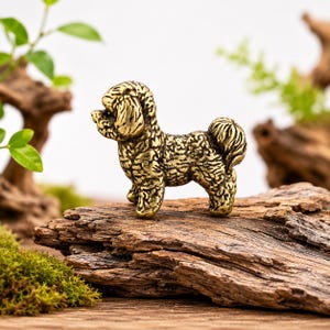 May include: A small, metallic gold-colored figurine of a dog with a curly coat, standing in profile. The sculpture features detailed, textured fur patterns across its body, a curled tail, and a distinct snout, capturing the likeness of a poodle.