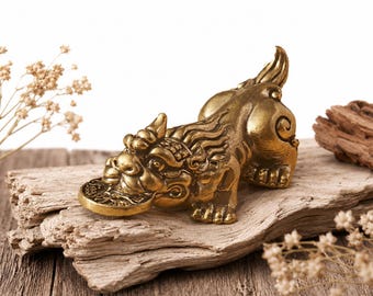 May include: A golden-colored, detailed figurine of a mythical creature, possibly a dragon or lion, holding a coin in its mouth. The figurine is posed on a piece of weathered wood, with dried flowers and wood pieces in the background.
