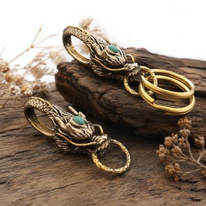 May include: Two brass dragon-shaped key clips featuring intricate scale textures and a small, smooth, oval-shaped turquoise-coloured stone set into the forehead of each dragon head. Each clip includes a circular brass ring for attaching keys.