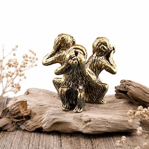 Peut inclure: Trois figurines de singes couleur laiton dans la pose « Ne rien voir, ne rien entendre, ne rien dire ». Les singes sont assis sur un morceau de bois flotté, avec des plantes séchées et des morceaux de bois en arrière-plan. Les figurines sont détaillées.
