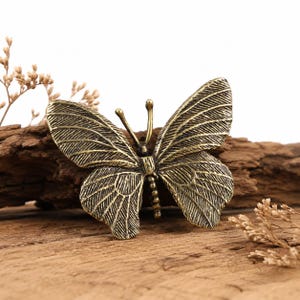 May include: A metallic butterfly-shaped object with an antique brass finish, featuring detailed engraved wing patterns, two thin antennae, and a segmented body. The butterfly is positioned with its wings spread wide, showcasing a textured surface.