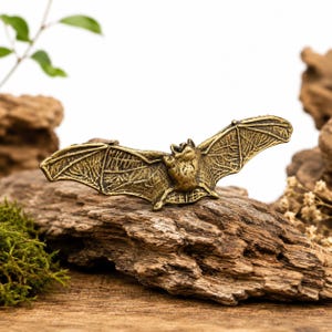 Puede incluir: Una pequeña figura de murciélago de metal color latón con las alas extendidas. El murciélago presenta membranas alares texturizadas y detalladas, además de un cuerpo esculpido, capturado en una pose de vuelo con las alas abiertas.