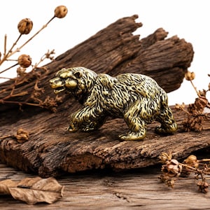 May include: A detailed brass bear figurine with textured fur, posed in a walking stance. The bear is set on a piece of weathered wood, with dried foliage accents. The figurine has a golden hue and is a decorative item.