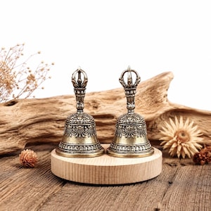 May include: Two antique bronze bells with ornate detailing, featuring a decorative handle and intricate patterns. The bells are displayed on a round wooden stand, set against a backdrop of driftwood and dried flowers. The bells have a vintage aesthetic.