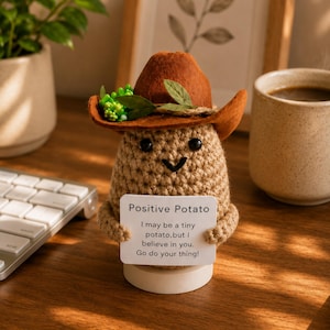 Puede incluir: Una figura de patata tejida a ganchillo con un sombrero de vaquero marrón con detalles verdes. La patata sostiene un cartel que dice "Positive Potato" con un mensaje inspirador. La figura está sobre una base blanca.