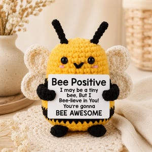 May include: A yellow and black crocheted bee plush toy holding a white rectangular sign that reads: Bee Positive, I may be a tiny bee, But I Bee-lieve in You! You're gonna BEE AWESOME. The bee has two black antennae, two cream-coloured wings, and black feet.
