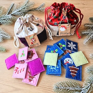 Peut inclure: Deux pochettes en tissu à cordon avec des motifs de Noël et des étiquettes en liège marquées Memory, accompagnées d'une collection de petits carrés en feutrine brodés de motifs festifs comme du houx, des flocons, des bonhommes de neige.