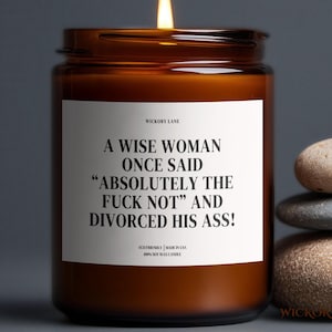May include: A lit brown glass candle jar with a white label that reads "A WISE WOMAN ONCE SAID 'ABSOLUTELY THE FUCK NOT' AND DIVORCED HIS ASS!". The candle is next to a stack of three stones.