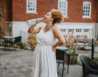 May include: A woman in a white maxi dress with a tiered skirt is smiling and drinking from a clear glass bottle. She is standing on a brick patio with chairs and tables in the background.