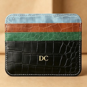 Może przedstawiać: Stos czterech skórzanych etui na karty w kolorze niebieskim, brązowym, zielonym i czarnym. Czarny etui ma złote inicjały "DC". Każdy etui ma fakturę skóry krokodyla i białe przeszycia.