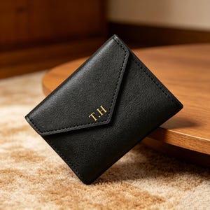 May include: A black envelope-style wallet with a textured finish and a pointed flap. The front of the wallet features the gold-colored embossed initials T.H. centered on the flap. The wallet has visible stitching along the edges.