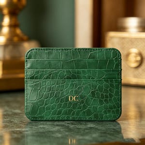 May include: A green cardholder with a crocodile skin texture. The cardholder has multiple card slots and the initials "DC" in gold lettering. The cardholder is rectangular and made of a textured material.