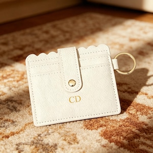 May include: A white card holder with a scalloped top edge, featuring a central vertical strap with a gold-toned snap button closure. The front is embossed with the letters CD in gold, and a gold-toned metal keyring is attached to the side.