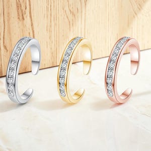 Può includere: Tre anelli aperti con incastonatura a binario e piccole pietre trasparenti a taglio rotondo. Il set include un anello color argento, uno color oro e uno color oro rosa, ciascuno con finitura lucida e un'apertura sulla fascia dell'anello.
