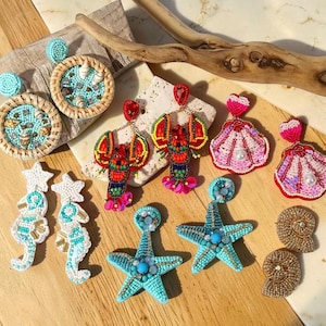 Op de afbeelding: Een collectie kralenoorbellen met diverse kustontwerpen, waaronder ronde gevlochten frames met turquoise kralen en schelpen, rode kreeftvormen, roze schelpvormen met parels, witte zeepaardjes, turquoise zeesterren en gouden spiraalschelpen.