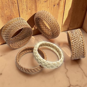 Op de afbeelding: Een verzameling van vijf ronde servetringen met een geweven rotan textuur. Vier van de ringen hebben een natuurlijke lichtbruine kleur, terwijl één ring in het midden een lichtere, gebroken witte kleur heeft, allemaal met een consistent vlechtpatroon.